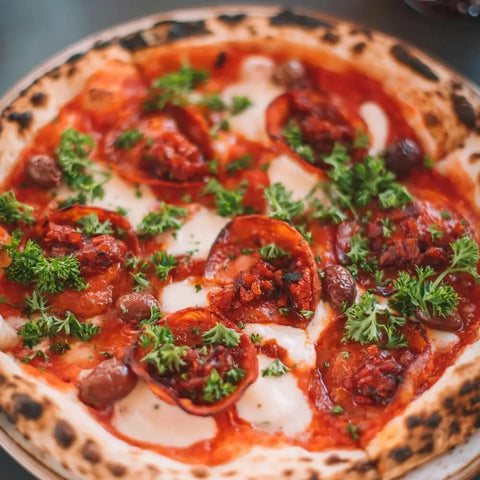 A close-up of a wood-fired pizza with tomato sauce, cheese, pepperoni, olives, and parsley on a golden crust, finished with a spicy kick from Gustus Vitaes Red Hot Sea Salts Collection - 3 Tins.