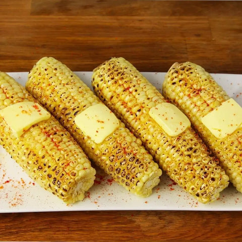 Four grilled corn cobs on a white plate, each topped with butter and sprinkled with Gustus Vitae Red Hot Sea Salts Collection for a smoky kick. The background features a wooden surface.