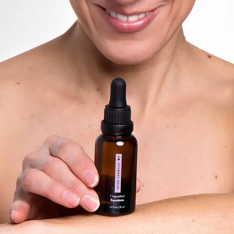 A smiling person holds a brown glass dropper bottle labeled Open Formula Squalane near their bare shoulder, showcasing how the Squalane moisturizer hydrates skin against a plain background.