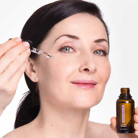 A woman with fair skin and dark hair applies Open Formula Squalane serum to her cheek with a dropper, holding the bottle in her hand. She smiles gently against a light background as the serum hydrates her skin.