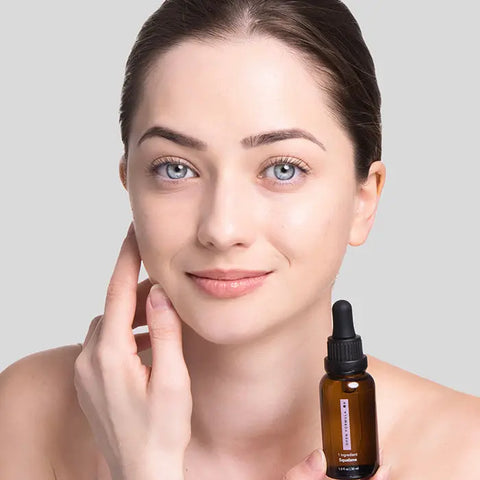 A woman with fair skin and blue eyes smiles gently, touching her cheek while holding an Open Formula Squalane dropper bottle, a moisturizing serum, against a plain light gray background.