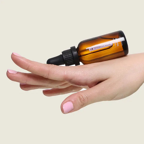 A hand with light pink nail polish balances an amber glass dropper bottle of Open Formula Squalane, a moisturizer for skin, horizontally across the fingers against a plain beige background.