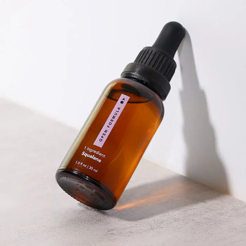 A brown glass dropper bottle labeled Open Formula Squalane 1.0 fl oz (30 ml) rests at an angle on a light surface, casting a shadow—ideal for moisturizing skin and protecting hair.