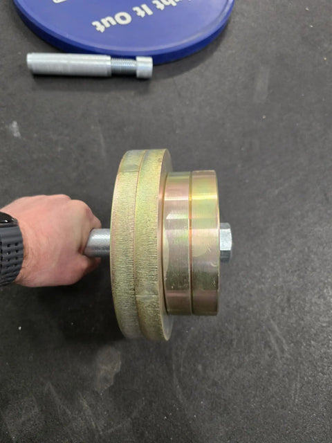 A hand grips a Weight It Out Dumbbell Standard 1 Handle Screwable Pair with two large plates secured by a nut. On the black gym floor, a blue plate and an Olympic plate adapter are nearby.