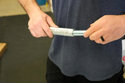 A person in a dark shirt and black ring holds a Weight It Out Dumbbell Standard 1 Handle Screwable Pair, gripping the threaded metal bar with an Olympic plate adapter labeled WEIGHT 1.0. The background is blurred.