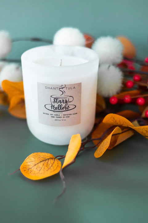 A white soy wax candle labeled Shanti + Tula Stars Hollow Inspired Soy Candle – Cozy Nostalgia Scent sits on a green surface, surrounded by yellow autumn leaves, red berries, and white fluffy decorations. Hand-poured in the USA.