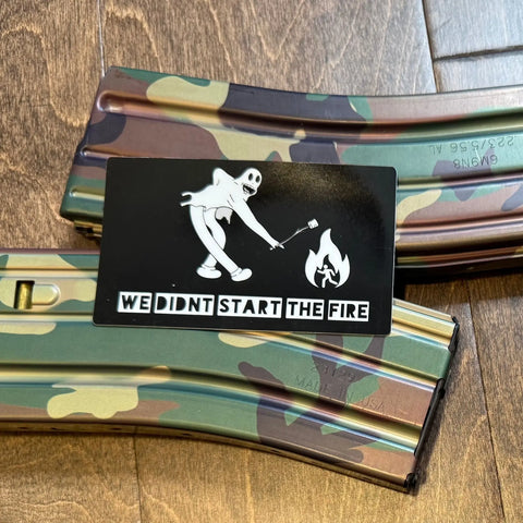 Two camouflage rifle magazines on a wooden floor with American Lictors Sticker: Start the Fire, featuring a skeleton roasting a marshmallow and the text WE DIDNT START THE FIRE—a truly unique design.