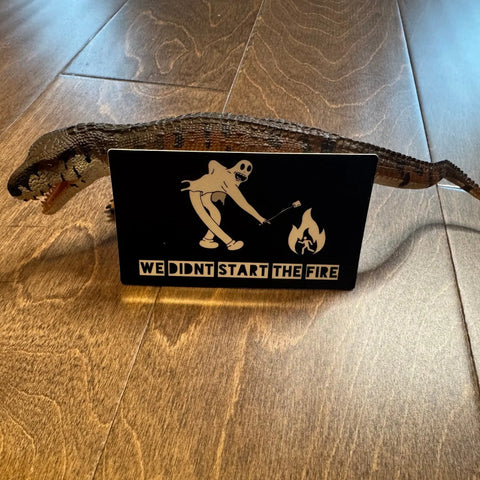 A toy dinosaur stands behind a 3x4 Sticker: Start the Fire by American Lictor, featuring a skeleton with a torch and flame and the text WE DIDNT START THE FIRE, displayed on a wooden floor. Designed by AGAVE.