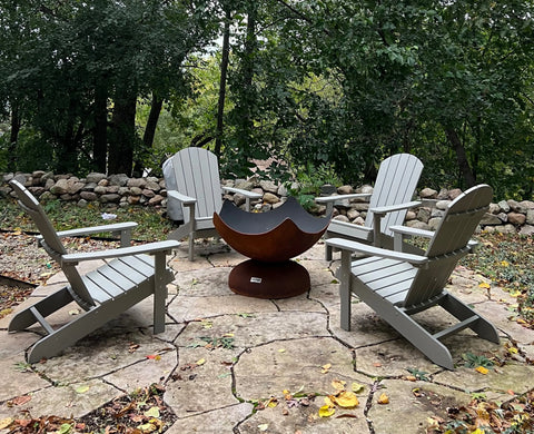 Four gray Adirondack chairs encircle the Stellar Artisan Fire Bowl by Ohio Flame on a stone patio, surrounded by trees and a rock wall, with fallen leaves scattered across the ground.