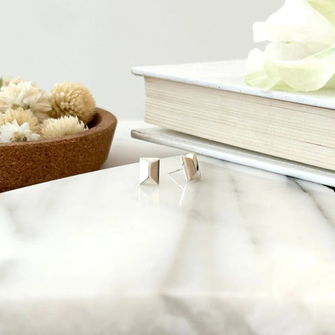 The SILVER CANAL STUDS by Sarah Cecelia are showcased on a white marble surface, with a closed book and a brown dish of dried flowers in the background, highlighting the elegance of these minimalist, handcrafted earrings.