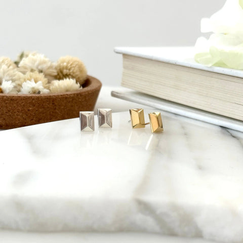 Two pairs of rectangular stud earrings—one gold, one handcrafted sterling silver (SILVER CANAL STUDS by Sarah Cecelia)—rest on a white marble surface, with a cork bowl of dried flowers and a closed book in the minimalist background.