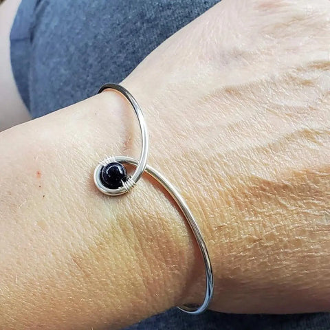 The Alexa Martha Designs Sterling Silver Blue Bead Ribbon Child Abuse Awareness Teardrop Bangle is shown wrapping a wrist, its open design featuring a teardrop with a blue bead at one end. A hand and grey clothing appear in the background.