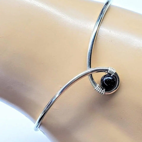 The Sterling Silver Blue Bead Ribbon Child Abuse Awareness Teardrop Bangle by Alexa Martha Designs features an open design with a single blue bead secured by wire wrapping, displayed on a mannequin wrist.