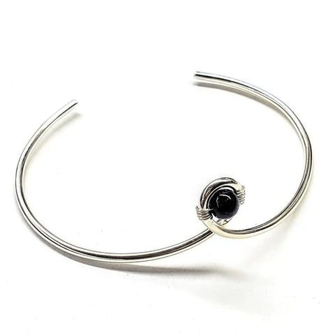 The Sterling Silver Blue Bead Ribbon Child Abuse Awareness Teardrop Bangle by Alexa Martha Designs features a minimalist open design with a blue bead centerpiece wrapped in silver wire.