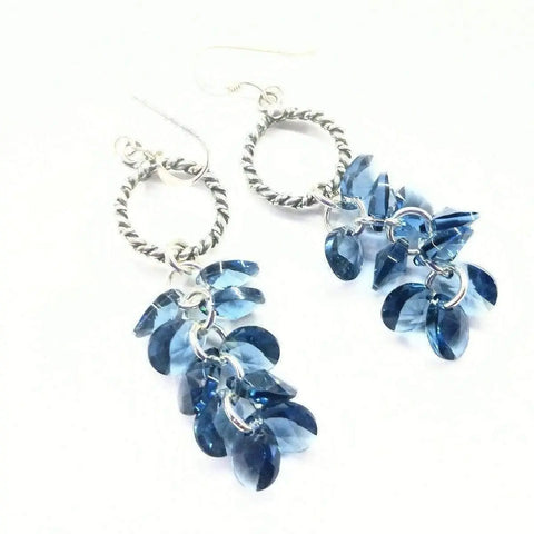The Sterling Silver Denim Blue Crystal Cluster Earrings by Alexa Martha Designs feature twisted circular hoops adorned with clusters of denim blue crystal beads. The Swarovski crystal earrings are shown on a white background.