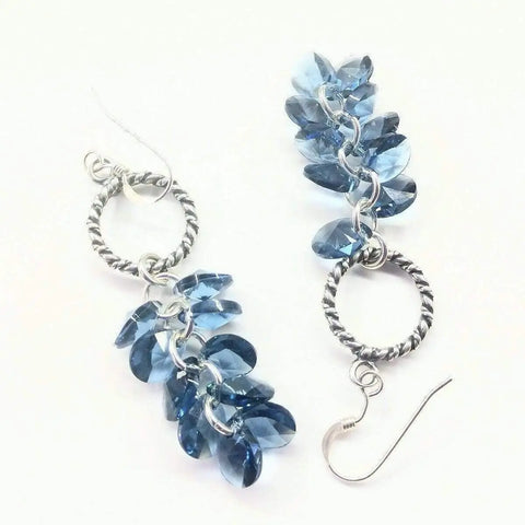 The Sterling Silver Denim Blue Crystal Cluster Earrings by Alexa Martha Designs feature twisted hoops with teardrop-shaped blue crystal beads, shown on a white background.