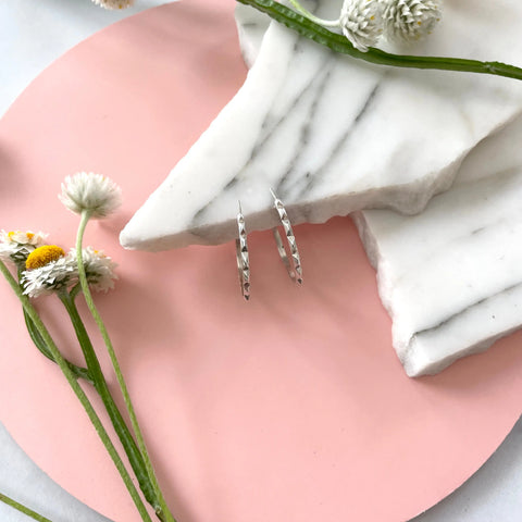 Sarah Cecelias SILVER CANAL HOOPS shown on stacked white marble slabs atop a pink circular surface, surrounded by scattered white and yellow flowers for decoration.