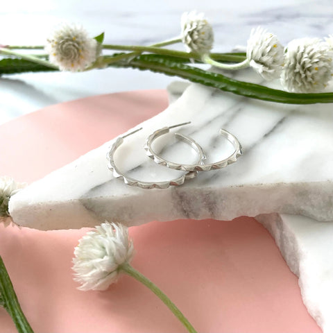 Sarah Cecelia SILVER CANAL HOOPS, handmade silver earrings with scalloped edges, are displayed on white marble among globe amaranth flowers and greenery, set against a pink and marble background.