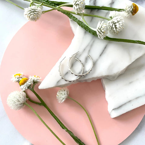 Sarah Cecelia’s SILVER CANAL HOOPS—elegant American-made sterling silver earrings displayed on white marble with dried flowers, set against a pink background—perfect for any occasion.