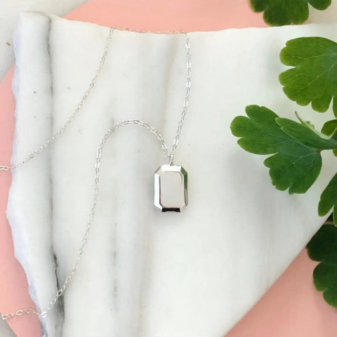 The SILVER GEMMA PENDANT by Sarah Cecelia, a minimalist silver layering necklace with a rectangular pendant, is displayed on white marble atop a pink surface with green leaves accenting the right side.