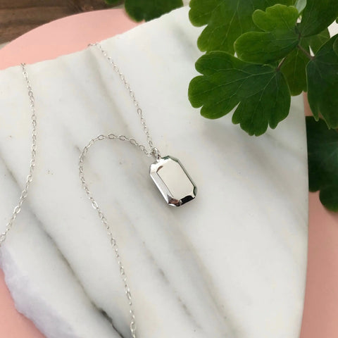 The SILVER GEMMA PENDANT by Sarah Cecelia—a minimalist rectangular silver necklace with beveled edges—rests on white marble, accented by green leaves and a hint of pink in the background.