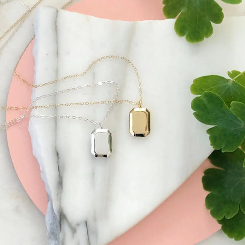 The SILVER GEMMA PENDANT by Sarah Cecelia, a rectangular sterling silver necklace, rests on a delicate chain over white marble and a pink background, with green leaves in the corner for a minimalist display.