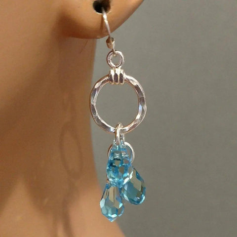 A close-up of Alexa Martha Designs Sterling Silver Hammered Aqua Crystal Cascading Drop Earrings, worn on an ear, showcases the handcrafted design and sparkling blue crystals against a neutral background.
