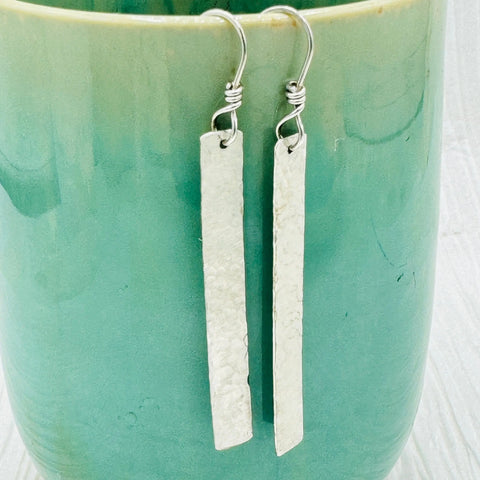 JANECKA Sterling Silver Hand Forged 2 Bar Earrings by Janecka, featuring wire-wrapped hooks, are displayed hanging on the rim of a glossy green ceramic cup.