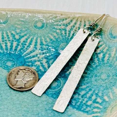 Janeckas JANECKA Sterling Silver Hand Forged 2 Bar Earrings, featuring a textured hammered finish, rest on a turquoise ceramic dish beside a U.S. dime to showcase their length and artisan craftsmanship.