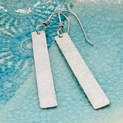 The Janecka Sterling Silver Hand Forged 2 Bar Earrings feature a rectangular, textured design with hook backs, and are shown on a blue textured surface.