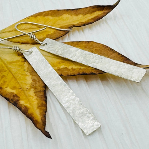JANECKA Sterling Silver Hand Forged 2 Bar Earrings by Janecka are displayed on yellow and brown dried leaves atop a light textured surface.
