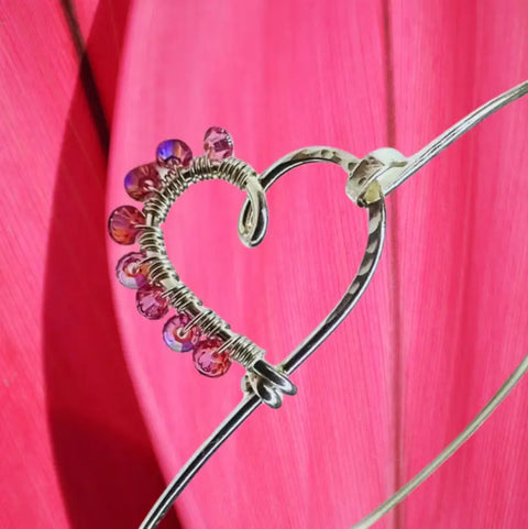 The Wire Wrapped Pink Crystal Heart Bangle by Alexa Martha Designs features a silver wire heart with pink and purple crystals, set against a bright, textured pink background.