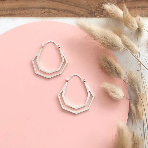 SILVER HERA HOOPS by Sarah Cecelia are displayed on a pink circular surface with beige dried grasses beside them and a wooden surface partly visible at the top. Handcrafted in the USA.