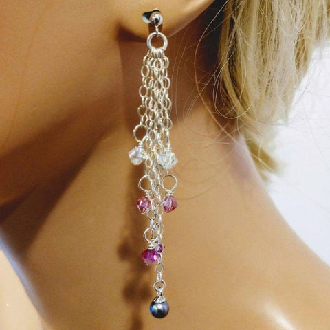 A close-up of Alexa Martha Designs’ Long Sterling Silver Crystal Pearl Tassel Earring, featuring silver chains, pink and clear beads, and a dark blue bead at the end, worn by a light-skinned person with blonde hair.