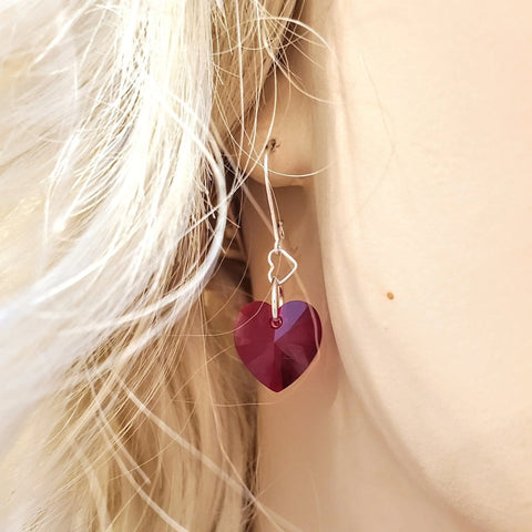 A close-up of a person’s ear with long blonde hair wearing Alexa Martha Designs’ Sterling Silver Red Heart Crystal I Love You Jesus Earrings, featuring handmade dangles with sterling silver and large, faceted red heart crystals.