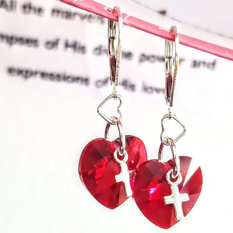 The Sterling Silver Red Heart Crystal I Love You Jesus Earrings by Alexa Martha Designs feature heart-shaped red pendants and silver cross charms on heart hooks, beautifully displayed on a pink rod.