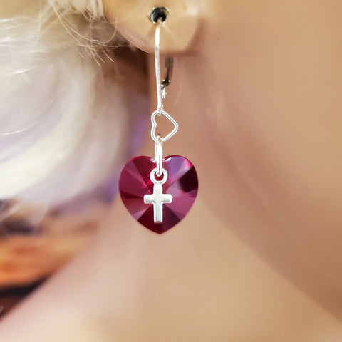 A close-up of an ear wearing Alexa Martha Designs Sterling Silver Red Heart Crystal I Love You Jesus Earrings, featuring a dangling red Swarovski heart charm and a small silver cross in the center.