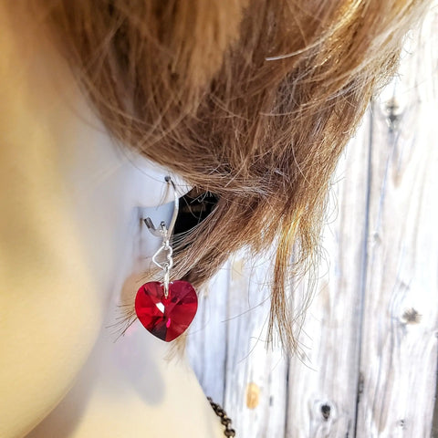 Close-up of a mannequin with short blonde hair wearing Alexa Martha Designs Sterling Silver Red Heart Crystal I Love You Jesus Earrings, featuring handmade dangles with shiny red crystals against a light wooden background.
