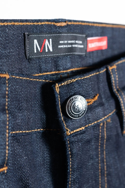 Close-up of The Nelson Dark Wash Denim by Marc Nelson shows dark blue 5-pocket jeans with a silver button, black waistband label featuring MN BY MARC NELSON in white and red, orange stitching, and a visible zipper fly.