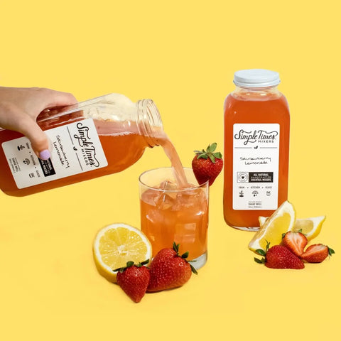 A hand pours Simple Times Mixers’ Strawberry Lemonade into a glass of ice as another bottle sits nearby with fresh strawberries and lemon wedges, perfect for cocktails, all set against a vibrant yellow background.