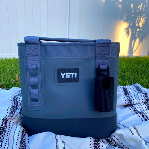 A Chatelet Manufacturing Sunglasses Holder Attachment is clipped to a gray YETI cooler with straps, resting on a blanket in the sunlight near a white fence—perfect for securely holding your sunglasses. Made in USA!.