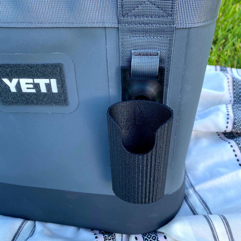 A gray YETI cooler sits on a blanket outdoors, featuring the Chatelet Manufacturing Sunglasses Holder Attachment—securely holding sunglasses on the cooler’s strap. Grass is visible in the background, showcasing this versatile accessory for coolers and backpacks.