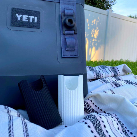 A black soft YETI cooler with a Chatelet Manufacturing Sunglasses Holder Attachment sits on a patterned blanket outdoors, with textured drink holders nearby. Green grass and a white fence are in the background.