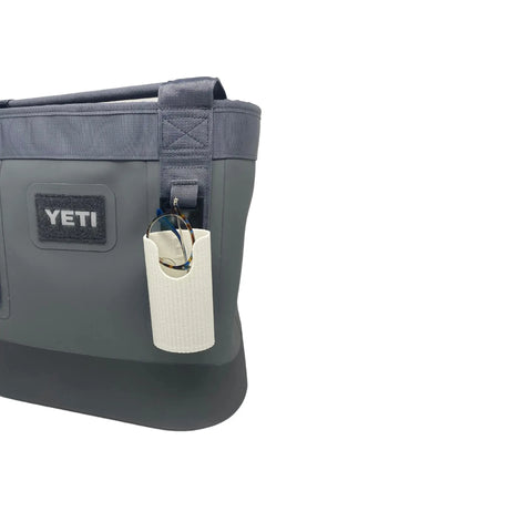A gray and black YETI bag features the Chatelet Manufacturing Sunglasses Holder Attachment, compatible with soft YETI coolers and backpacks. The textured, light-colored accessory is attached to its side strap securely holding a pair of sunglasses. White background.