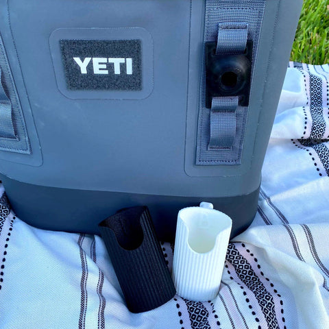 A gray Yeti cooler sits on a striped picnic blanket outdoors with two Chatelet Manufacturing Sunglasses Holder Attachments for Yeti coolers and backpacks displayed upright in front. Grass is visible in the background.