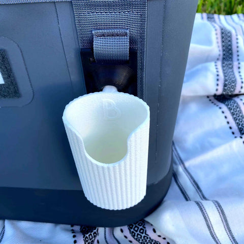 A Chatelet Manufacturing Sunglasses Holder Attachment for Soft Yeti Coolers & Backpacks is secured to a gray Yeti cooler, which sits on a patterned black and white cloth outdoors on the grass.