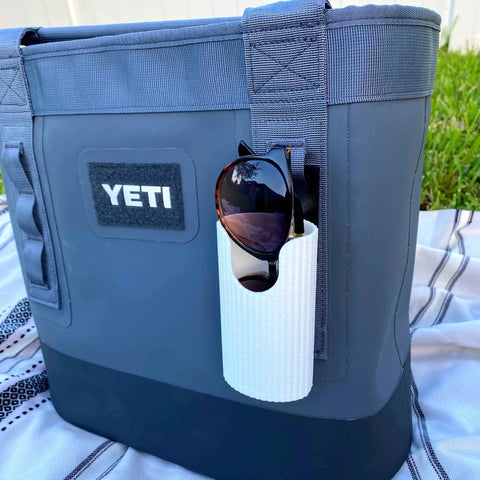 A Chatelet Manufacturing Sunglasses Holder Attachment, made in the USA for soft YETI coolers and backpacks with straps, secures a pair of sunglasses to a gray YETI cooler bag resting outdoors on a blanket with grass in the background.