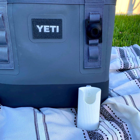 A Chatelet Manufacturing Sunglasses Holder Attachment is secured to a gray YETI cooler with straps, resting on a patterned blanket outdoors. A small white device lies in front, with green grass visible in the background.