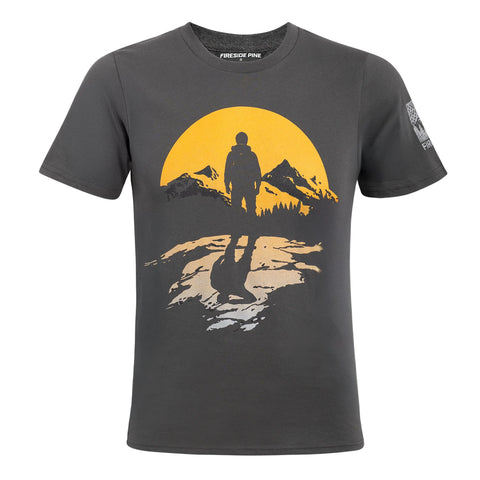 The Sunrise Hiker T-shirt by Fireside Pine features a graphic of a hiker on rocky ground, silhouetted against a yellow sun with mountains and trees. Made in the USA, this relaxed fit tee includes brand detail inside the collar.