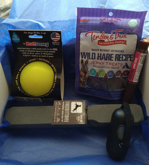 The Super Max Indestructibone Gift Box by Bulletproof Pet Products Inc for dogs 101 lbs+ features a yellow indestructible dog chew toy, Tender & True Wild Hare Recipe Jerky Treats, beef stick, waste bag dispenser, and bags on blue tissue paper.